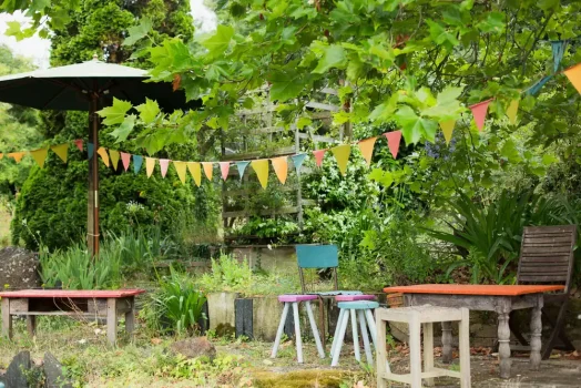 Alt : Jardin convivial avec mobilier coloré, parasol, guirlande de drapeaux et végétation luxuriante.