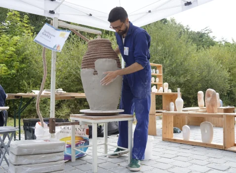 2. Artiste créant une sculpture en terre cuite lors d'un atelier en plein air avec plusieurs œuvres en cours autour de lui, sous un chalet en extérieur.